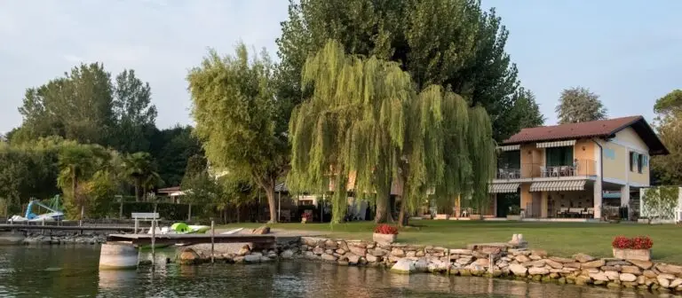 Una casa in riva al lago, con il tetto rosso e un ampio balcone, sorge dietro un muro di pietra e un prato rigoglioso, circondata da alberi ad alto fusto e da un salice piangente, con un piccolo pontile e una barca sulle acque tranquille in primo piano.