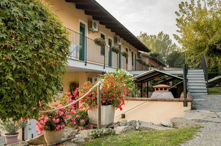 Un edificio giallo a due piani con un balcone adornato da vasi di piante, scale esterne, una vegetazione rigogliosa e fiori colorati in primo piano, in una giornata tranquilla.