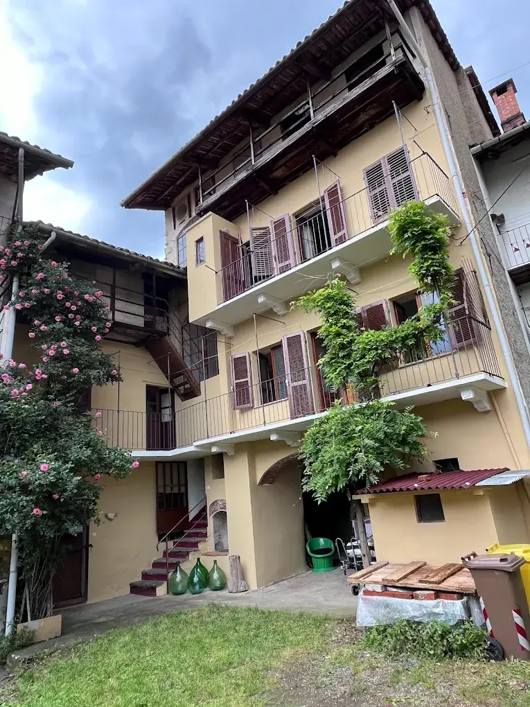 Next to a lush flowering bush stands a yellow multi-storey building, with shutters and wooden balconies, partly covered with green climbing. In the courtyard you can see various objects, including bottles and bins.