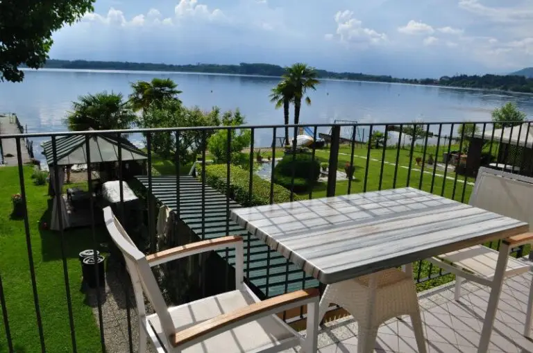 Un balcone con un tavolino e delle sedie si affaccia su un giardino con palme, un gazebo e un laghetto, sotto un cielo parzialmente nuvoloso.