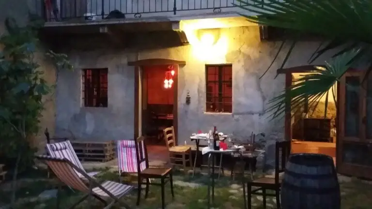 A cozy outdoor patio with wooden chairs, a table set for dinner and a barrel, in front of a rustic building lit by soft lights in the evening. In the foreground you can see a lush plant.