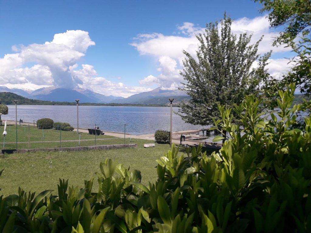Una vista panoramica su un lago con rive erbose, alberi e montagne sullo sfondo, sotto un cielo azzurro punteggiato di nuvole. Dei lampioni costeggiano un sentiero lungo la riva, mentre in primo piano si vedono piante rigogliose.
