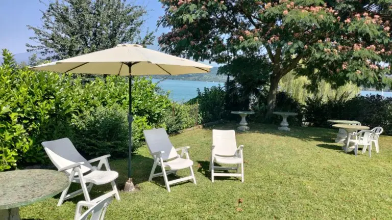 Un giardino soleggiato con sedie a sdraio bianche, un grande ombrellone, tavolini e alberi rigogliosi. Sullo sfondo, si intravede uno specchio d'acqua oltre a fitti cespugli e fogliame.