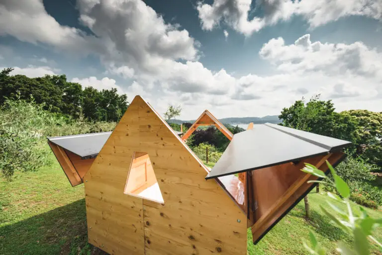 Una piccola e moderna casetta di legno dalle pareti triangolari e con i lati aperti sorge su un prato, circondata da alberi, sotto un cielo parzialmente nuvoloso.