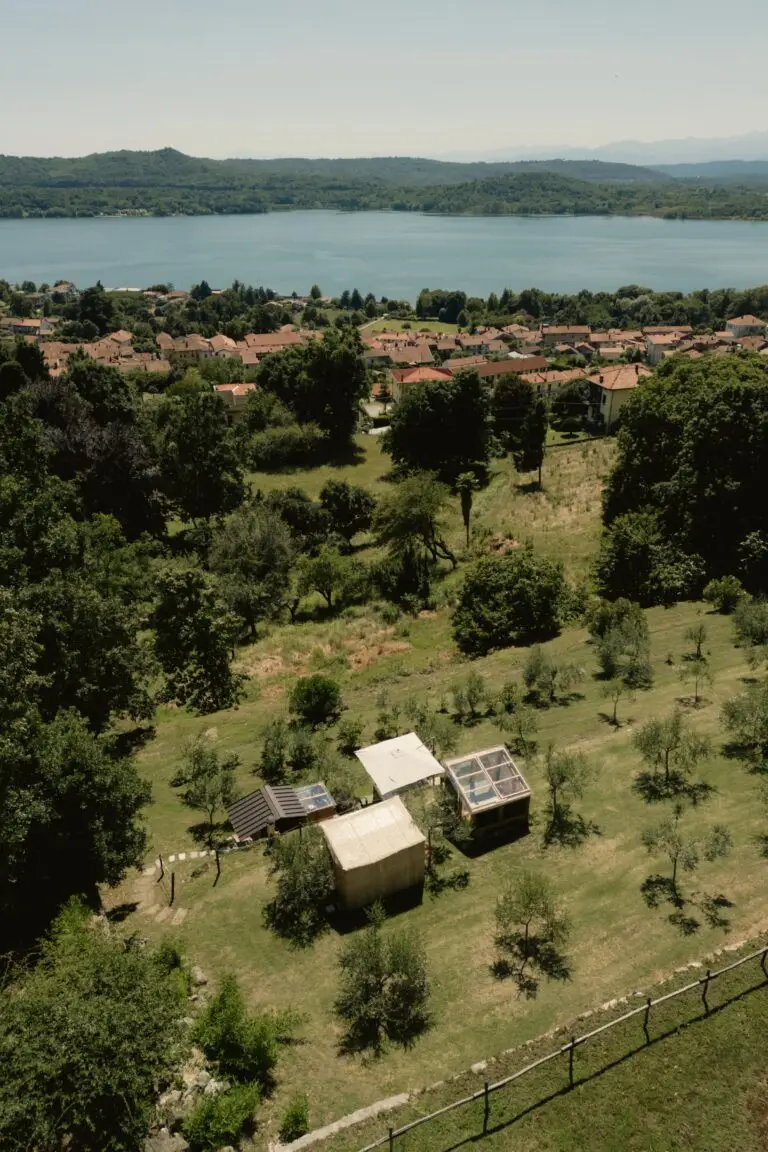 Vista aerea di un paesaggio erboso con piccoli capanni, alberi sparsi, un villaggio con case dai tetti rossi e, sullo sfondo, un grande lago circondato da verdi colline sotto un cielo limpido.