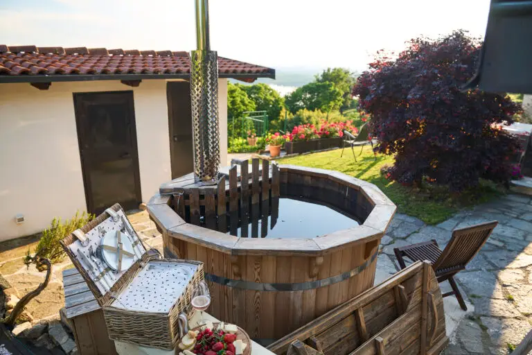 Una vasca idromassaggio in legno è collocata su una terrazza in pietra accanto a una casa, circondata dal verde e da piante in fiore. Nelle vicinanze c'è un cestino da picnic aperto con piatti e frutti di bosco, mentre alcune sedie in legno sono disposte di fronte alla vista sul giardino.