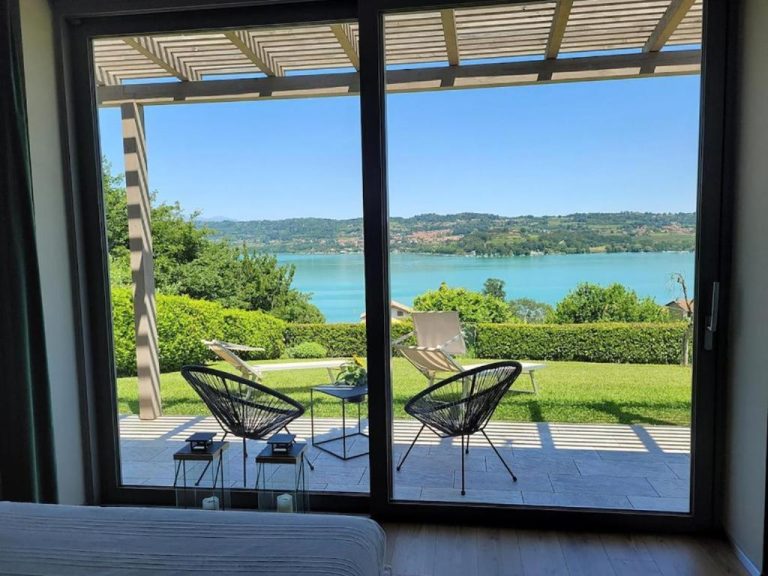 Looking through the large glass doors you see a patio with two modern chairs and a coffee table, overlooking a green lawn, a blue lake and hills in the distance under a clear sky.