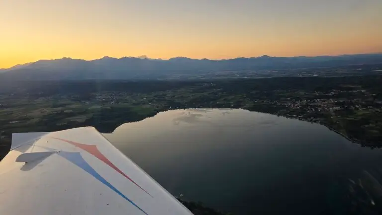 Aerial view of a quiet lake surrounded by greenery and small villages at sunset, with a mountain range in the background and a part of the wing of an airplane visible in the foreground.