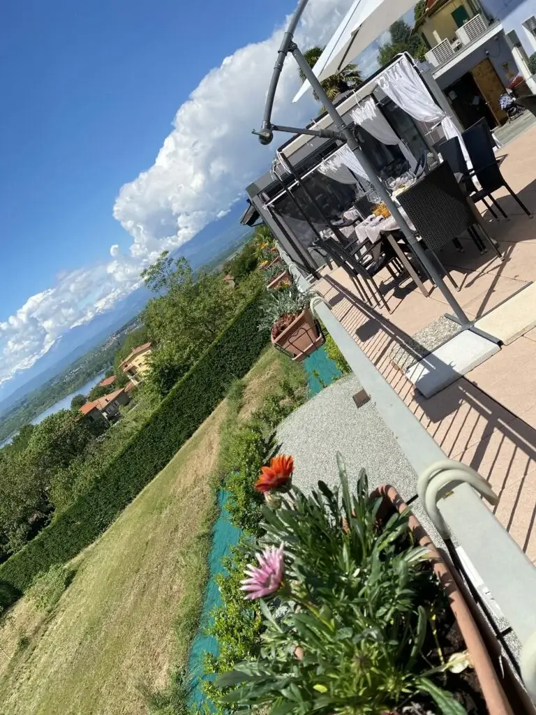 Un balcone arredato con mobili da esterno e tende bianche si affaccia su un giardino verdeggiante con vasi di fiori in primo piano. La vista comprende alberi, case vicine e montagne in lontananza sotto un cielo parzialmente nuvoloso.