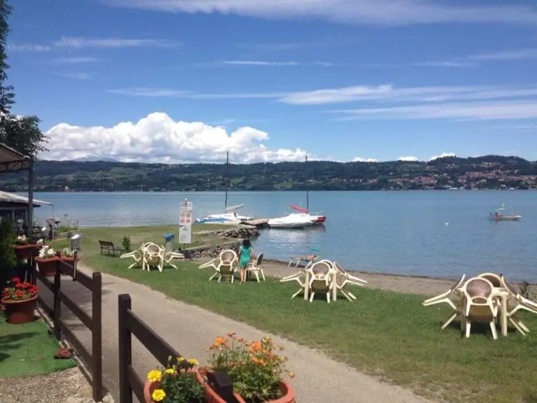 A scene by the lake with plastic chairs and tables on a lawn, boats moored along the shore, a small path, flower pots and hills in the background, under a partially cloudy sky.