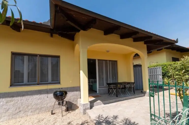 Casa gialla a un piano con veranda ombreggiata, tavolo e sedie da giardino, un piccolo barbecue nero e un vialetto pavimentato, circondata dal verde e da una recinzione, sotto un cielo azzurro e limpido.
