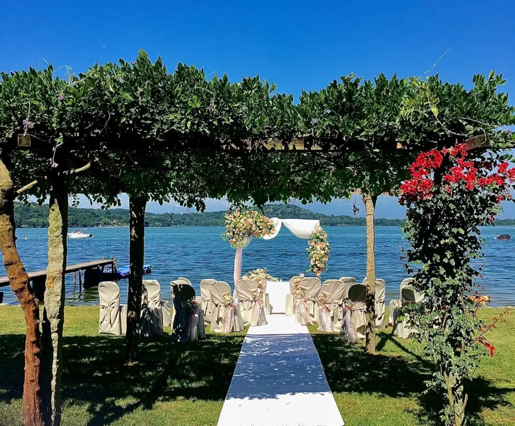 L'allestimento per un matrimonio in riva al lago prevede un tappeto bianco che conduce a un arco adornato di fiori sotto un pergolato ricoperto di vite. Le file di sedie, decorate con fodere bianche e fiocchi, fanno da cornice alle acque blu e al cielo limpido.