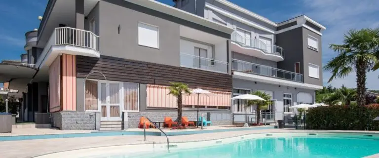A modern grey building with balconies and large windows overlooking a pool with crystal clear waters, surrounded by palm trees, deck chairs and umbrellas on a sunny day.