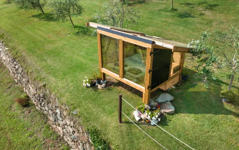 Una piccola serra in legno e vetro sorge su una collinetta erbosa accanto a un muro di pietra, circondata da piante e fiori, con alberi sparsi sullo sfondo.