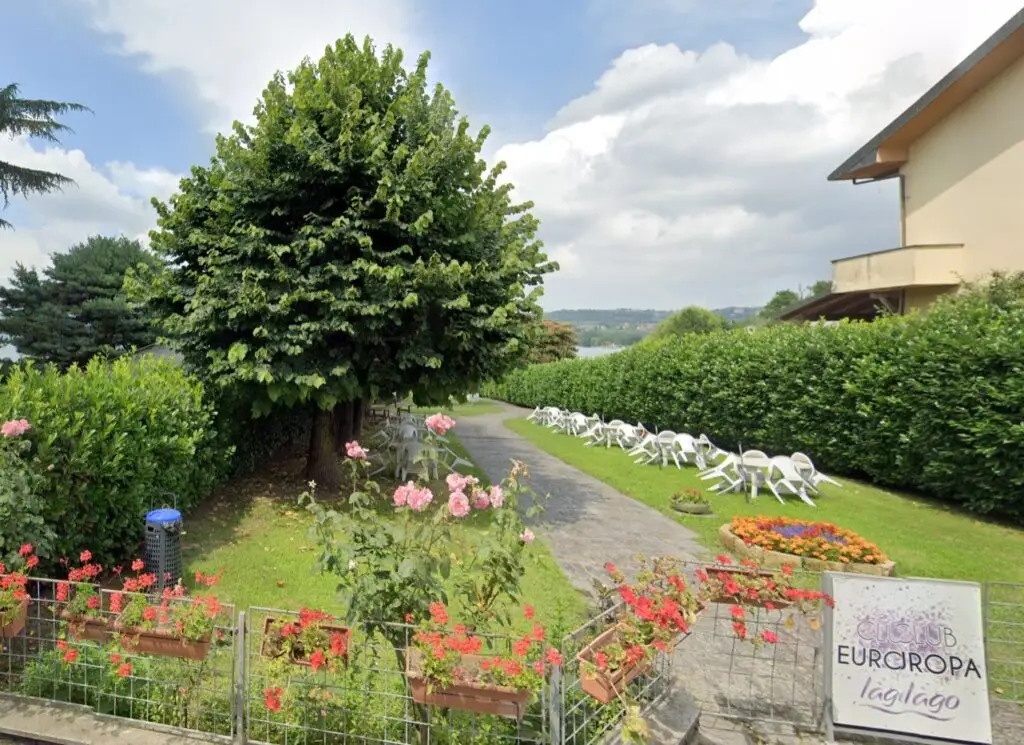 Un sentiero nel giardino, fiancheggiato da alberi, fiori e siepi, conduce verso un lago in lontananza; sul prato sono disposte delle sedie a sdraio bianche e un cartello recita: «Camping Europa Il lago».