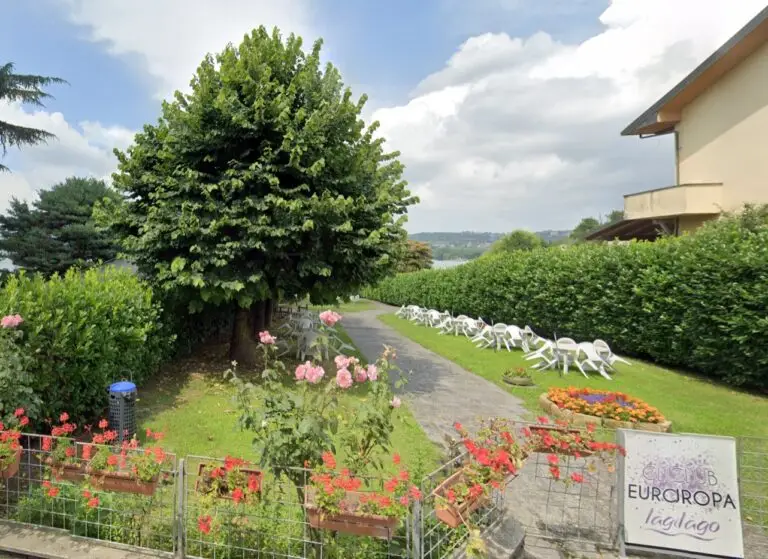 Un sentiero nel giardino, fiancheggiato da alberi, fiori e siepi, conduce verso un lago in lontananza; sul prato sono disposte delle sedie a sdraio bianche e un cartello recita: «Camping Europa Il lago».