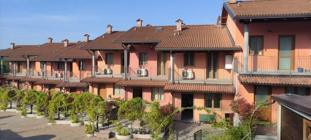 Una fila di villette a schiera arancioni e marroni con balconi e tetti spioventi, dotate di unità esterne di climatizzazione. Sotto, un cortile con piante in vaso, rampicanti e alberelli sotto un cielo azzurro e limpido.