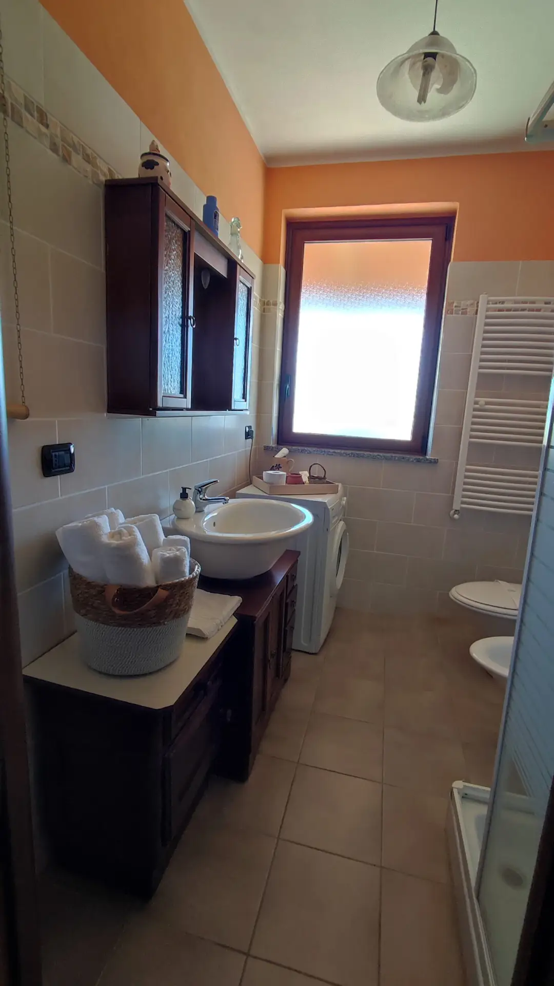 Un bagno stretto con piastrelle beige, un lavandino con specchio, una lavatrice, un WC, un bidet, una doccia, un scaldasalviette e un cesto di asciugamani appoggiato su una cassettiera di legno. La luce del sole filtra da una finestra.