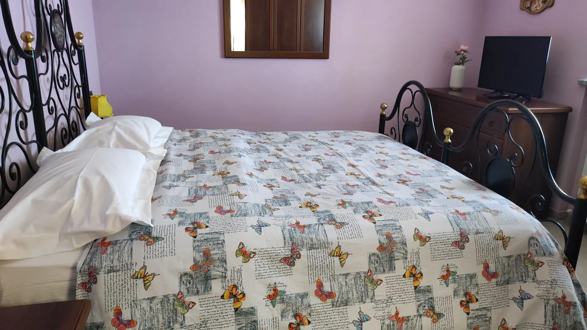 Un letto ben rifatto con un copriletto decorato con farfalle e motivi a caratteri, cuscini bianchi e una testiera in metallo nero lavorata. Su una parete color viola chiaro è appeso un comò in legno decorato con uno specchio.
