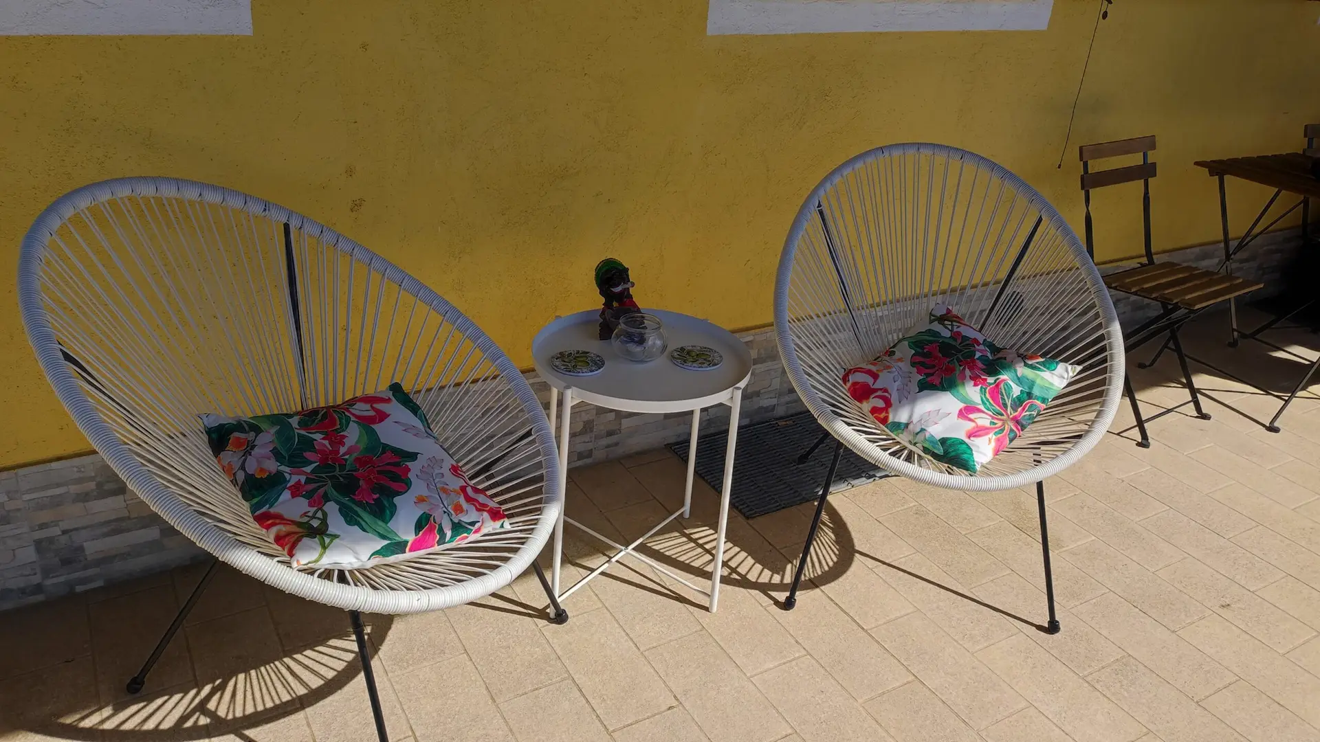 Due sedie bianche di forma circolare con cuscini a motivi floreali sono disposte su un patio piastrellato accanto a un tavolino rotondo su cui sono appoggiati una bambola, un bicchiere e due piatti. Lo scenario fa da sfondo a una parete gialla, con una sedia di legno nelle vicinanze.
