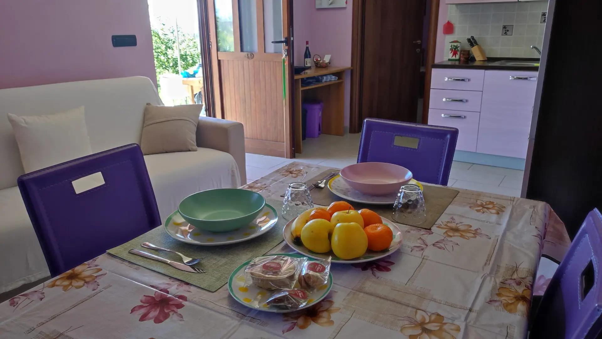 Un tavolo da pranzo apparecchiato per due con ciotole colorate, bicchieri, posate, frutta assortita e pasticcini. La stanza comprende un divano, una porta aperta sul giardino e una cucina con mobili color lavanda.