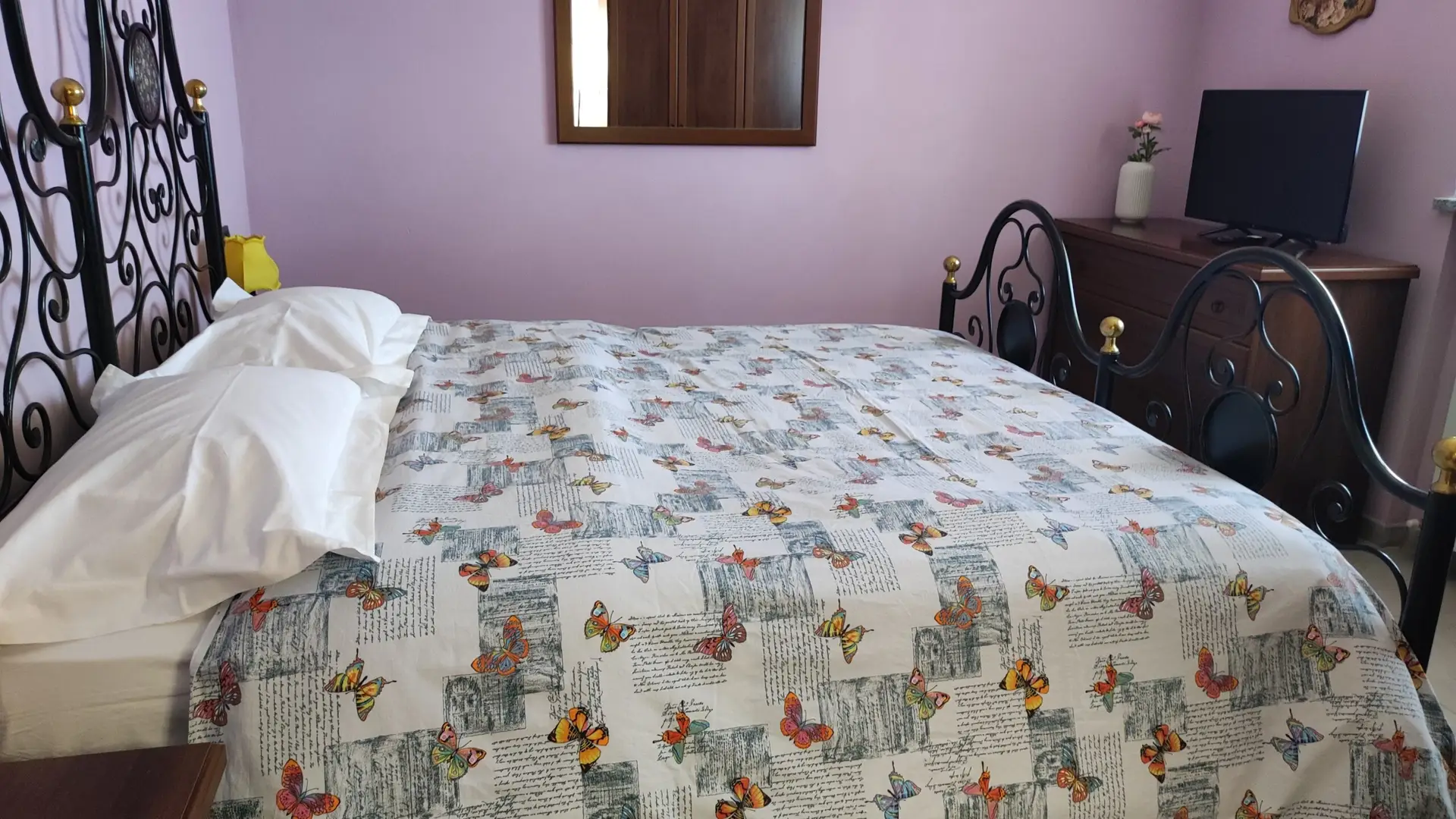 Un letto ben rifatto con un copriletto decorato con motivi a farfalle e scritte, due cuscini bianchi e una testiera in metallo lavorata. Un comò con specchio, vaso e oggetti decorativi è addossato a una parete color lavanda.