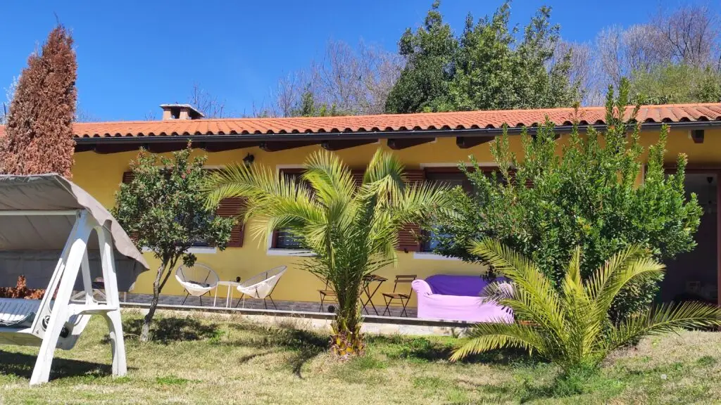 Una casa gialla a un piano con il tetto di tegole rosse, circondata da una vegetazione rigogliosa e da palme. Davanti, in una giornata di sole, ci sono un'altalena, sedie da giardino e un divano da esterno rivestito di tessuto viola.