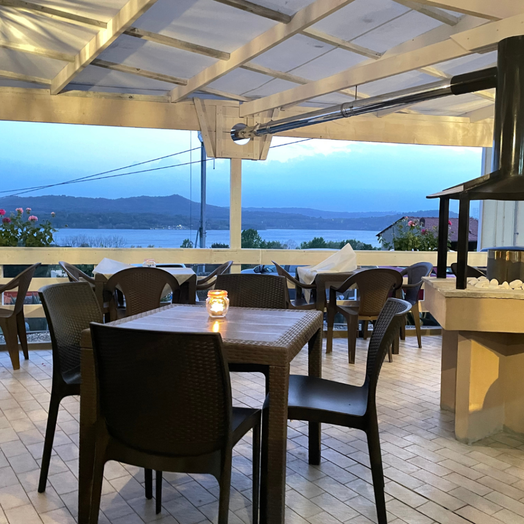 Terrazza di un ristorante all'aperto con tavoli e sedie neri, una candela accesa su un tavolo e una vista sul mare e sulle montagne al tramonto. L'area è coperta da un pergolato bianco e decorata con vasi di fiori lungo i bordi.