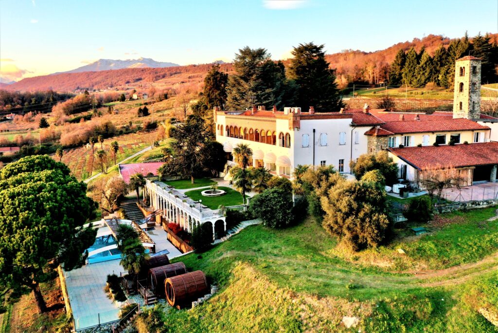 Una grande villa con balconi ad arco e tetto di tegole rosse sorge su una collina, circondata da vigneti e alberi, con le montagne sullo sfondo e una piscina e una terrazza in primo piano.