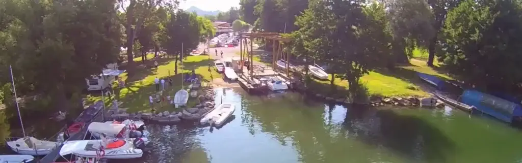 Un porto turistico in riva al lago con barche ormeggiate lungo la riva, circondato da alberi verdeggianti e prati. Si vedono diverse persone e automobili vicino all'acqua, mentre sullo sfondo si intravedono alcuni piccoli edifici.