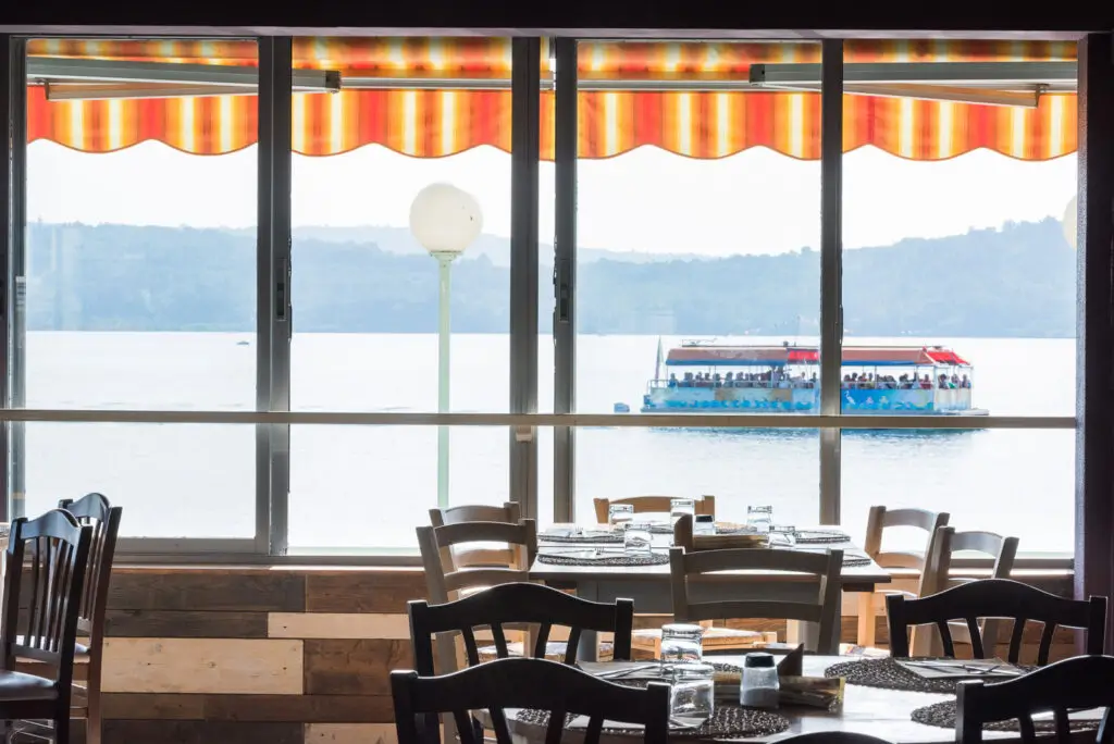 Dall'interno si vede un ristorante sul lago con i tavoli apparecchiati. Fuori dalla finestra, una barca con delle persone a bordo galleggia sul lago sotto una tenda da sole a strisce arancioni, con le colline visibili in lontananza.