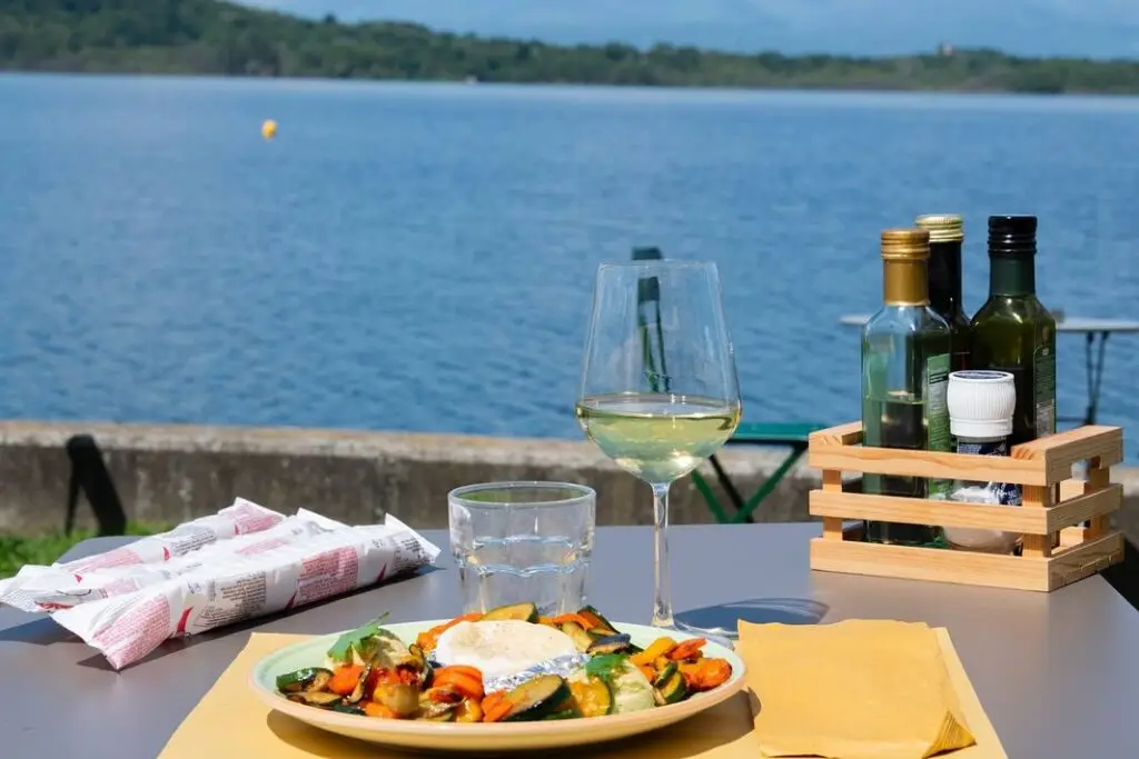 Su un tavolo all'aperto in riva al lago sono disposti un piatto di cibo e un bicchiere di vino bianco, con accanto alcune bottiglie di olio d'oliva e alcuni condimenti. Sullo sfondo si vedono le acque tranquille e le verdi colline ricoperte di alberi.