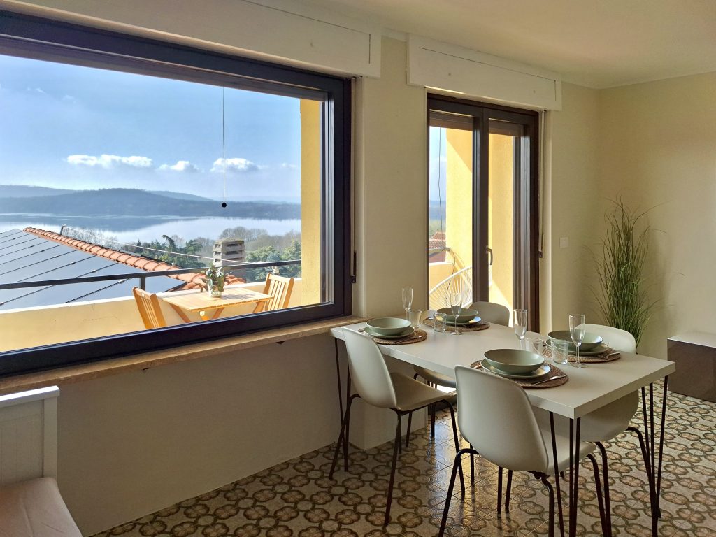 Una luminosa sala da pranzo con un tavolo apparecchiato per quattro persone, sedie bianche e ampie finestre che offrono una vista panoramica sulle montagne, sul lago e su un patio esterno con tavolo e sedie. La luce naturale inonda la stanza.