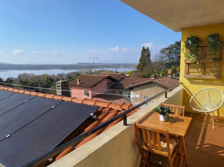 Un balcone soleggiato, arredato con un tavolo di legno, sedie, piante in vaso e fioriere a muro, si affaccia su case dai tetti rossi, un grande lago e alberi in lontananza, il tutto sotto un cielo azzurro e limpido.