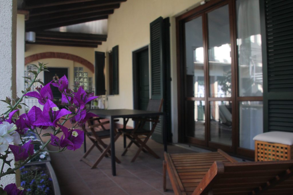 Un patio coperto con sedie e tavolo in legno, persiane verdi, porte a vetri e, in primo piano, una fioriera con vivaci fiori viola.