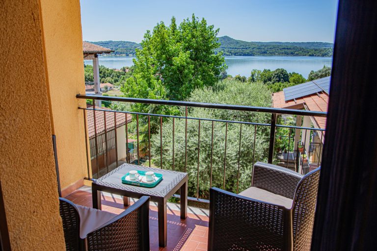 A small balcony with two wicker chairs and a coffee table overlooks the rooftops, green trees and a lake with hills in the distance, under a clear blue sky.