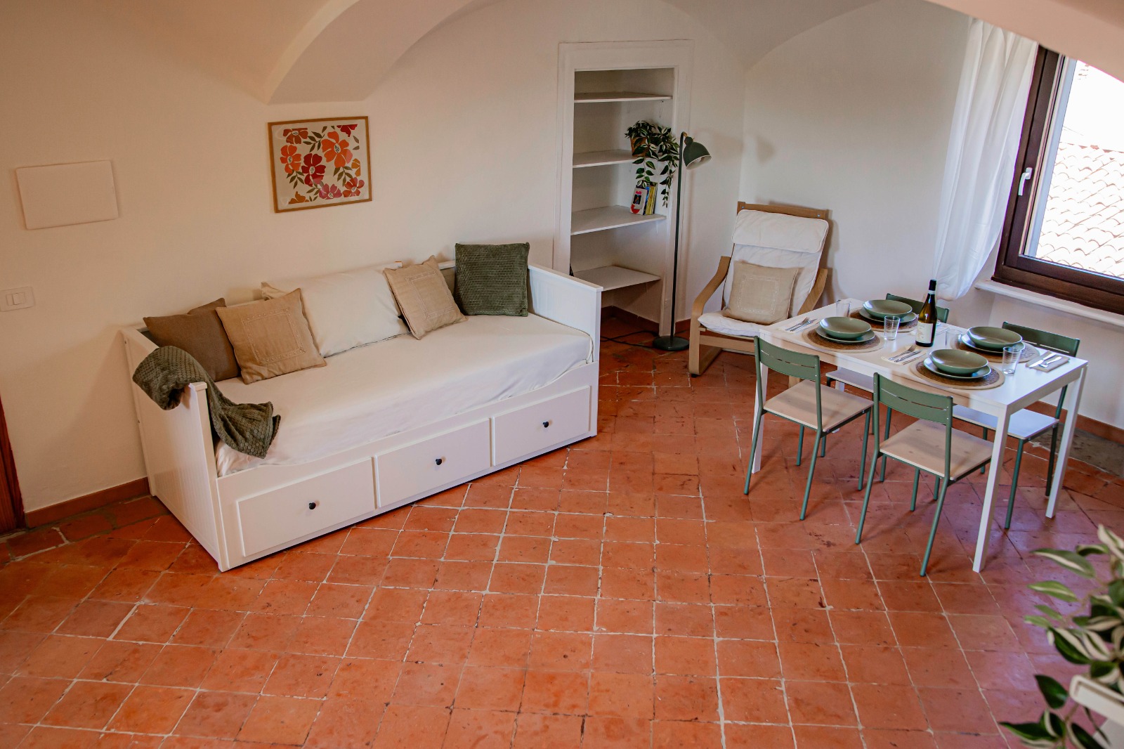 Un accogliente salottino con un divano letto bianco ricoperto di cuscini, una libreria, una poltrona e un tavolino da pranzo per due persone vicino alla finestra, il tutto su un pavimento in cotto.