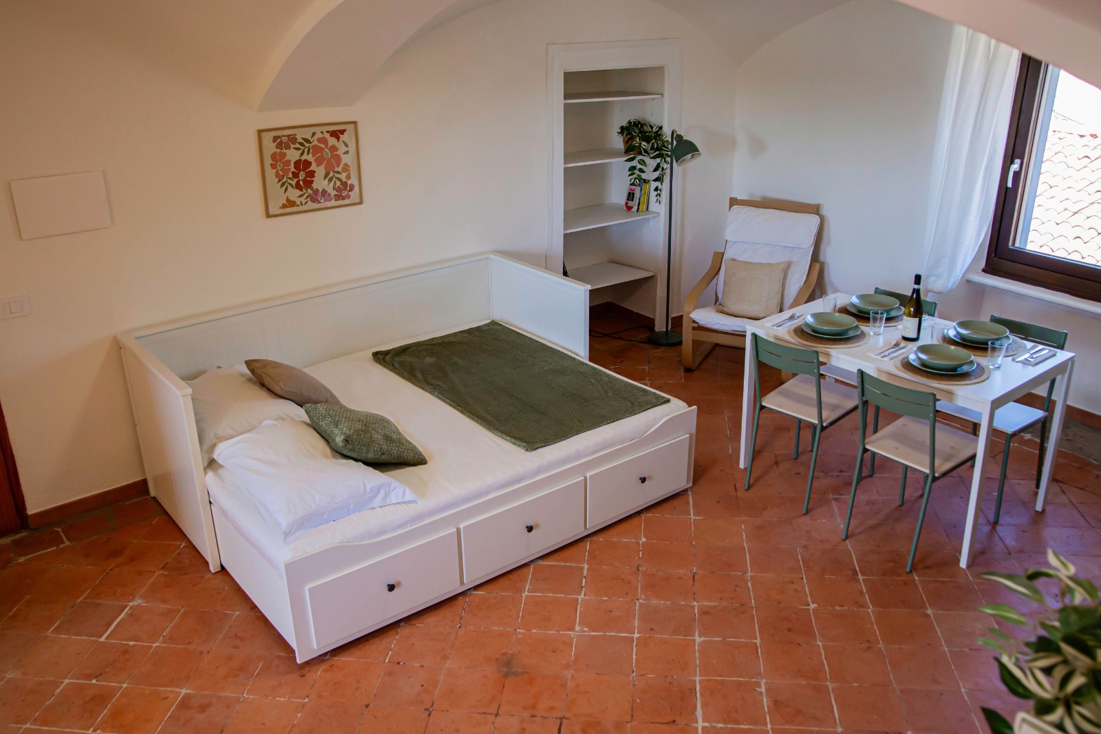Una stanza accogliente con un letto bianco dotato di cassetti, un piccolo tavolo da pranzo per quattro persone, una sedia in legno con un cuscino, una libreria bianca con delle piante e una finestra con tende bianche. Il pavimento è in cotto.