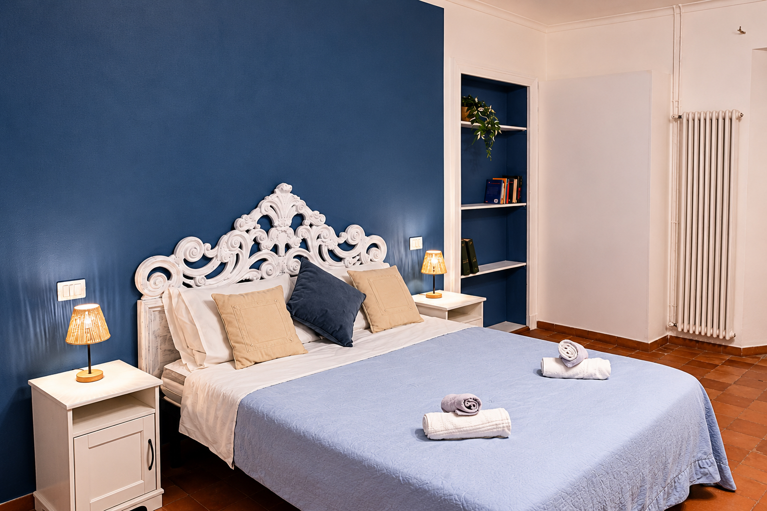 Un letto ben rifatto, con una testiera bianca decorativa, fa da contrappunto a una parete blu. Due comodini con lampade affiancano il letto, sul quale sono appoggiati degli asciugamani piegati. Nella stanza, accogliente e ordinata, si intravedono delle mensole e un termosifone.