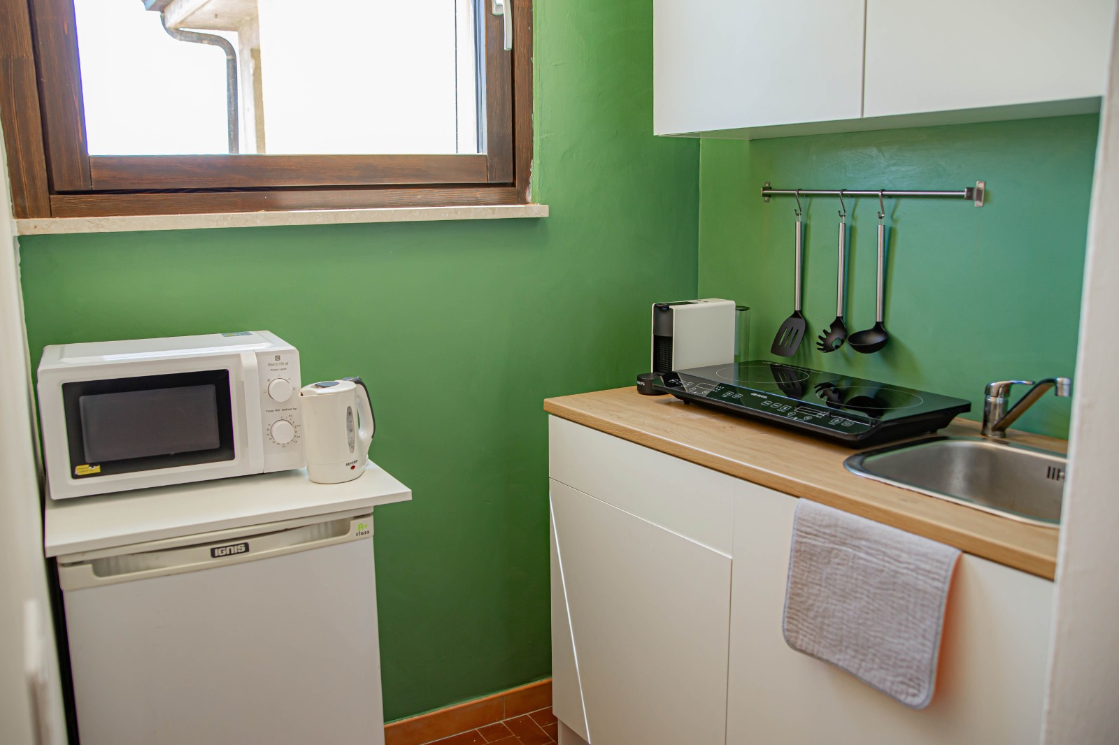 Una piccola cucina con pareti verdi, dotata di forno a microonde e bollitore elettrico appoggiati su un mini-frigo, un piano cottura con utensili appesi sopra, un lavello, mobili bianchi e un asciugamano appeso a un cassetto. Una finestra lascia entrare la luce naturale.