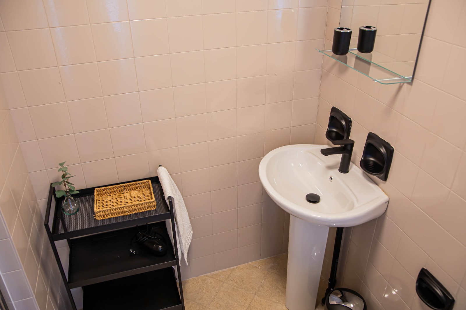Un piccolo angolo bagno con un lavabo a colonna bianco, rubinetteria nera, una mensola in vetro con due tazze nere e un carrello nero su cui sono appoggiati un vassoio di vimini, una pianta e un asciugamano. Pareti e pavimento rivestiti di piastrelle beige.