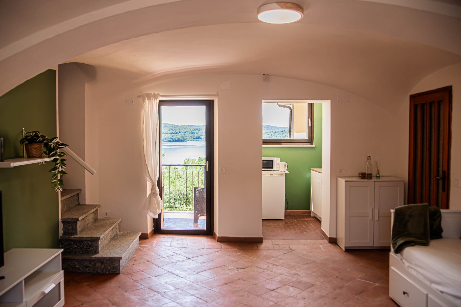 Una stanza luminosa con pavimenti in cotto, pareti verdi e bianche, una piccola scala, un mobile bianco e una porta che conduce a un balcone con vista sul mare e sulle colline. Attraverso una porta aperta si intravede un angolo cottura.