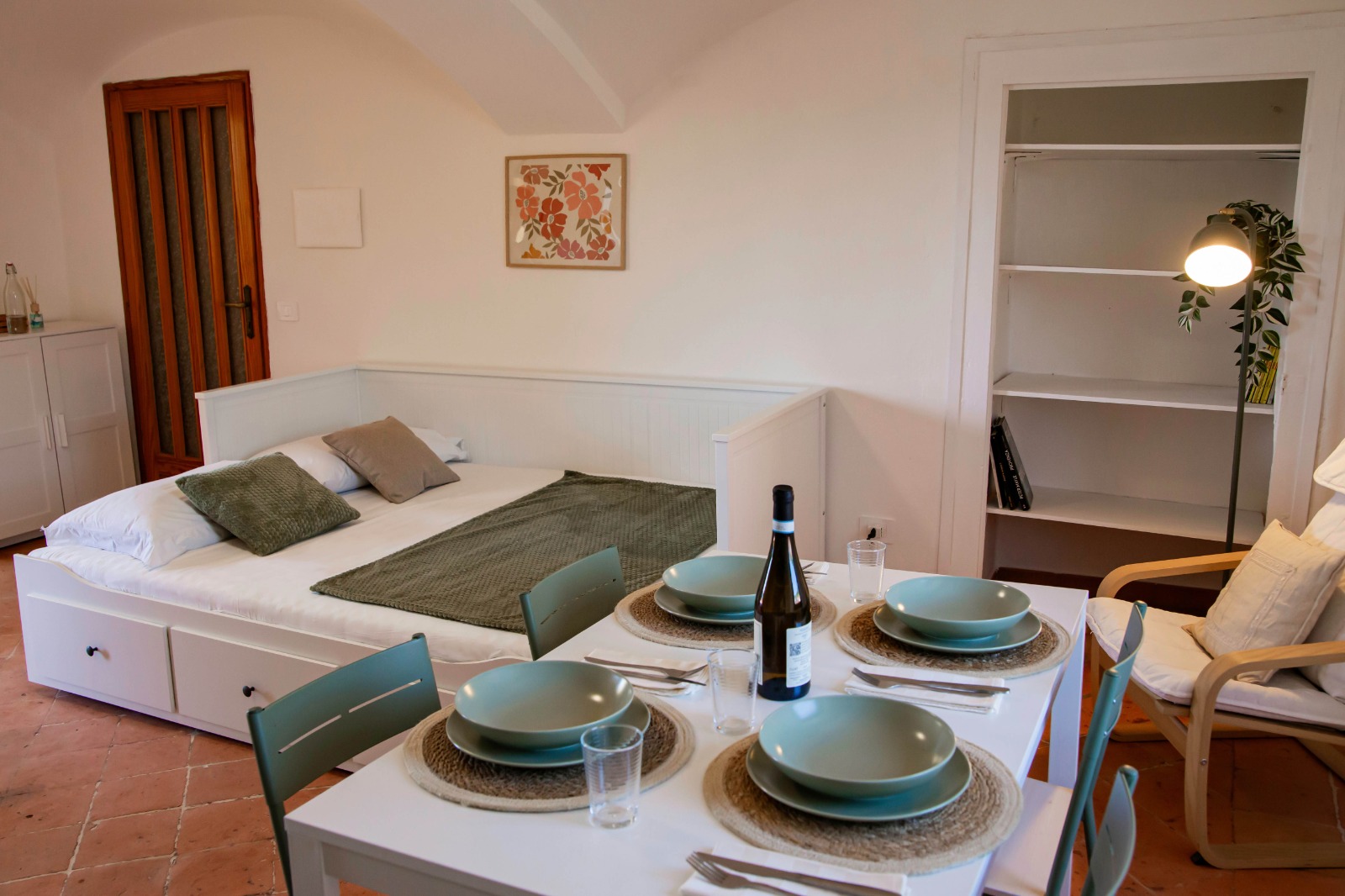 Un monolocale accogliente con un letto, tocchi di verde, un tavolino da pranzo per quattro persone, una poltrona con una lampada e una libreria a vista. La stanza è luminosa e invitante, con un arredamento semplice.