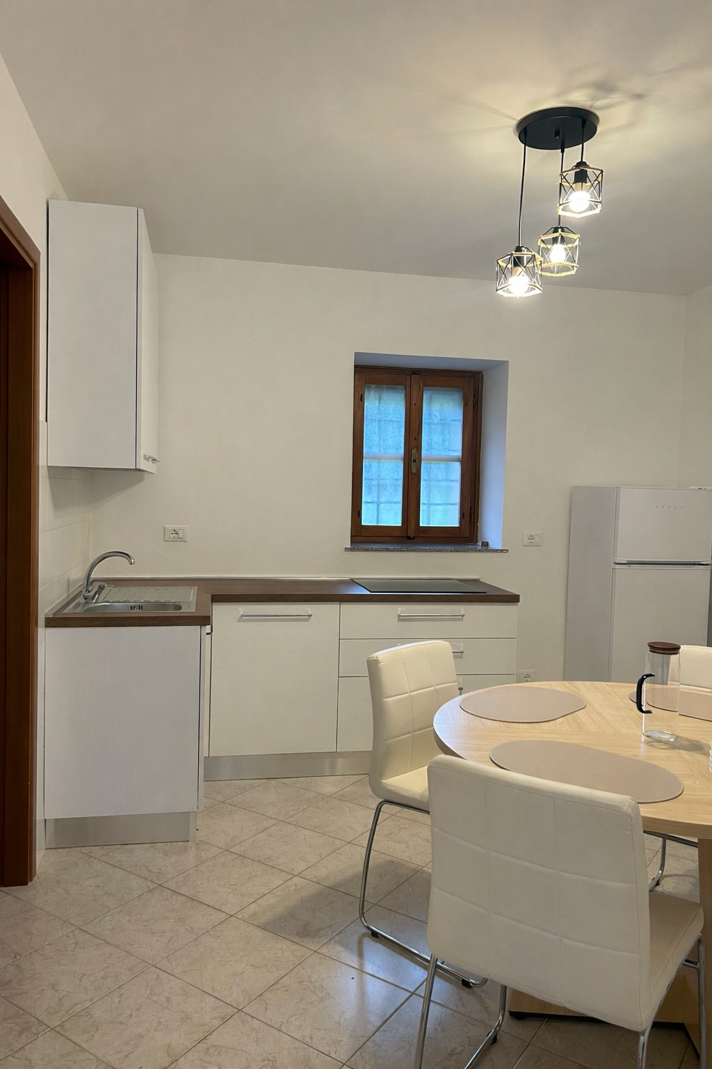 Una cucina moderna con mobili bianchi, un tavolo da pranzo rotondo con quattro sedie bianche, un piccolo lavello, un frigorifero, una finestra con infissi in legno e una lampada a soffitto a tre luci.