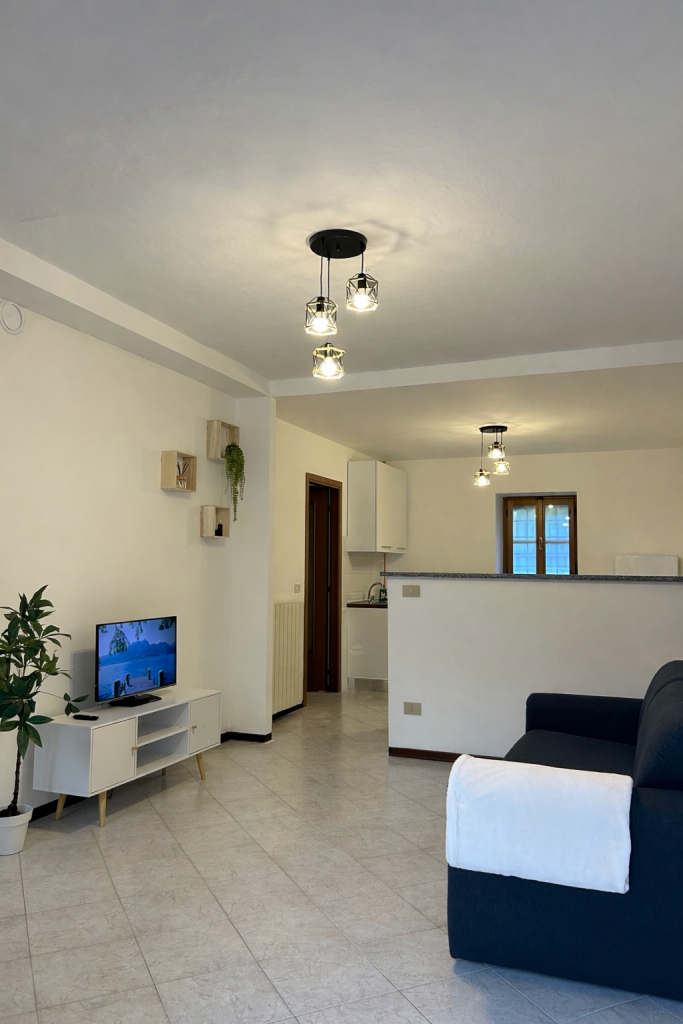 Un soggiorno moderno con un divano blu scuro, una TV su un mobile bianco, una pianta in vaso, mensole a muro e lampade a soffitto. Lo spazio conduce a una zona cucina con una finestra e un arredamento minimalista.