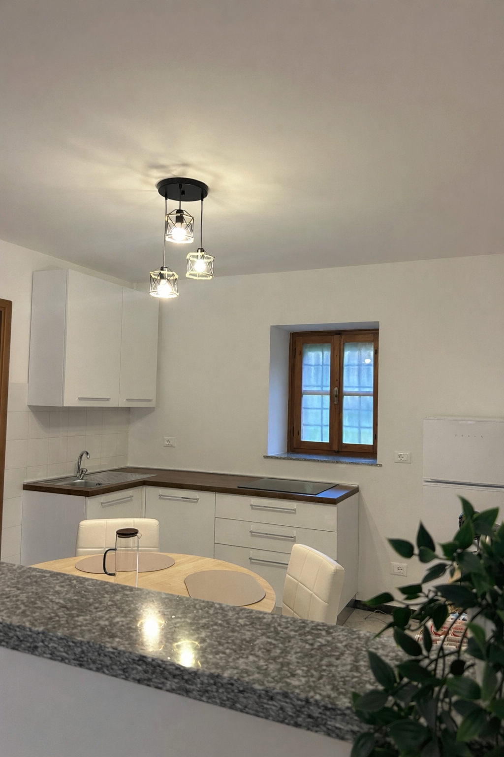 Cucina moderna con mobili bianchi, un piano di lavoro in legno, un tavolino rotondo con sedie bianche, una tazza di ceramica, una finestra con infissi in legno, un frigorifero e una pianta in primo piano sotto le luci a soffitto.