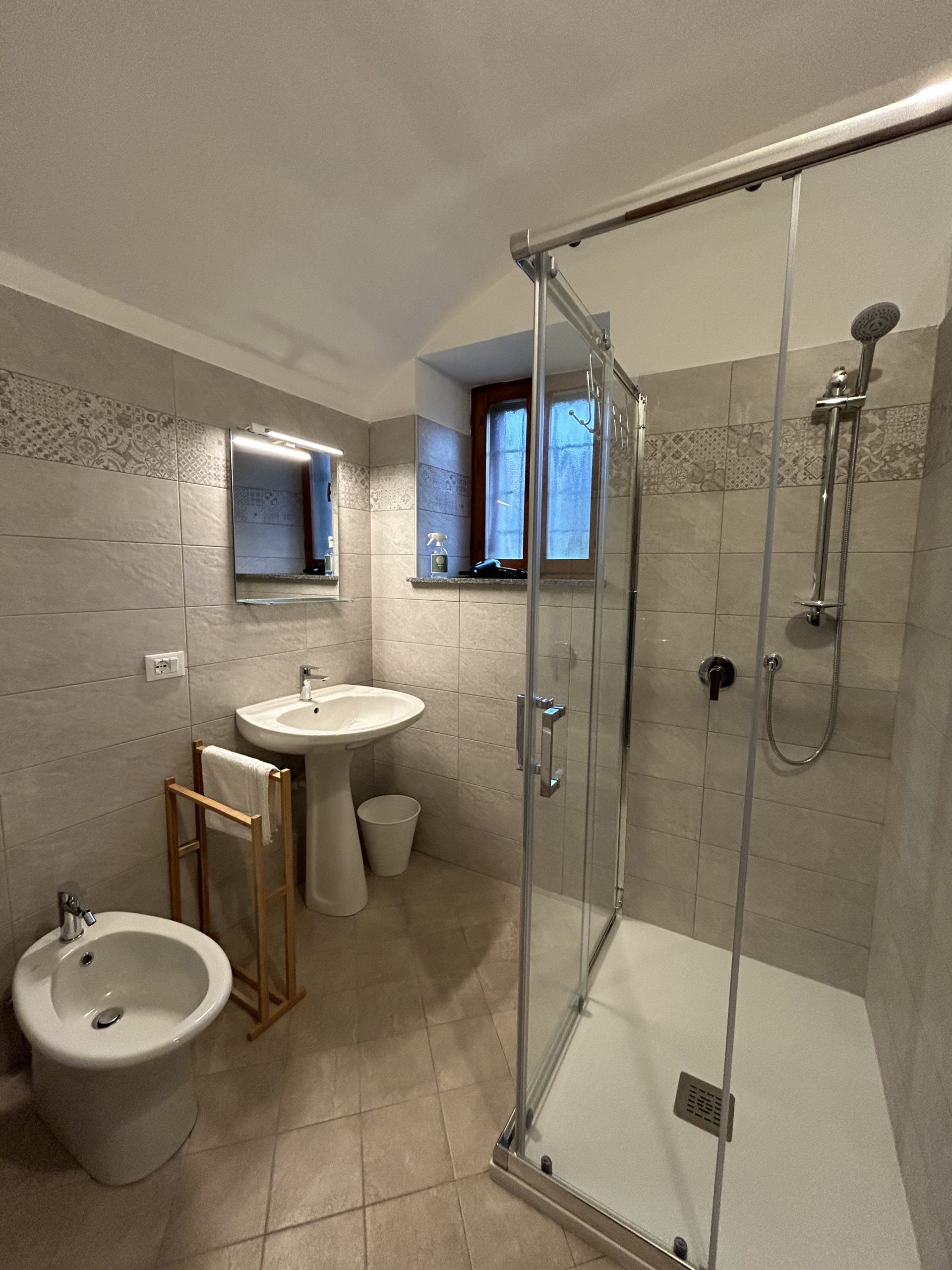 Bagno moderno con pareti e pavimento piastrellati in beige, dotato di box doccia in vetro, lavabo su piedistallo con specchio, bidet, piccolo cestino dei rifiuti bianco, portasciugamani in legno e una finestra che lascia entrare la luce naturale.