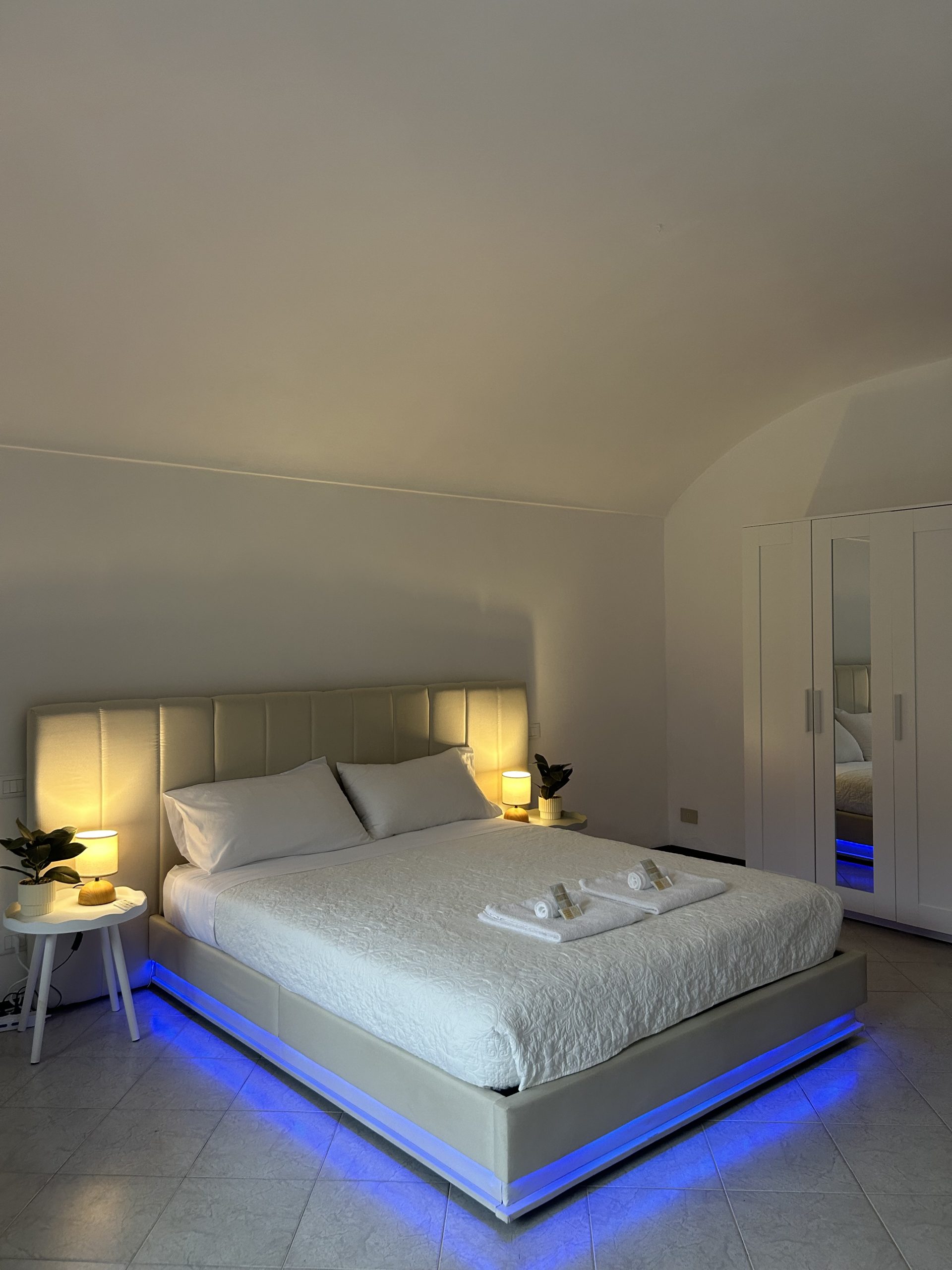 Una camera da letto moderna con un grande letto con biancheria da letto bianca e luci LED blu sotto il materasso, due comodini con lampade e piante, e un armadio bianco con ante a specchio addossato a una parete liscia.