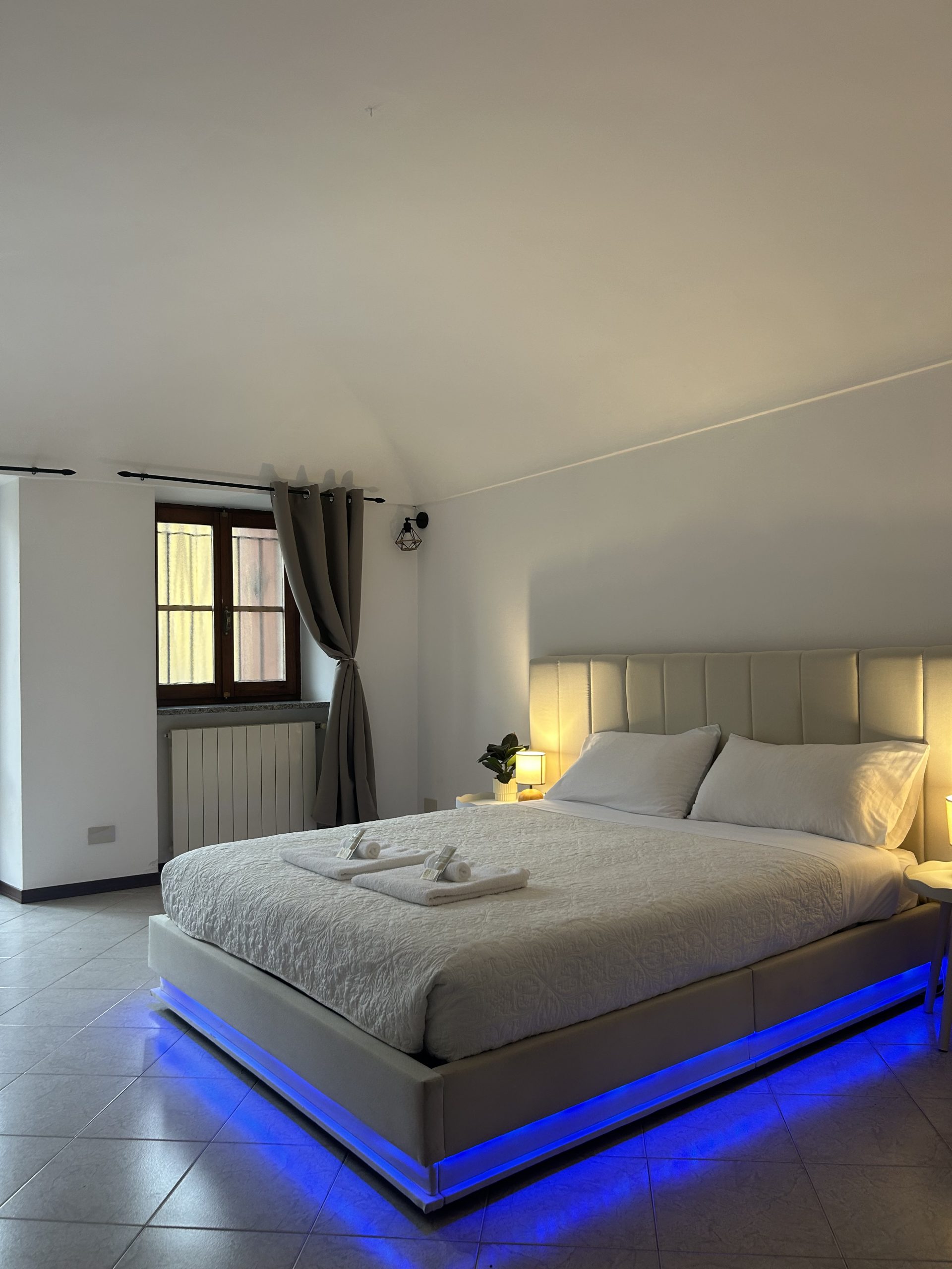Camera da letto moderna con un grande letto con biancheria da letto bianca e luci LED blu sotto il materasso, due asciugamani piegati con cura sul letto, una finestra con tende grigie e una piccola lampada da comodino su un tavolino.