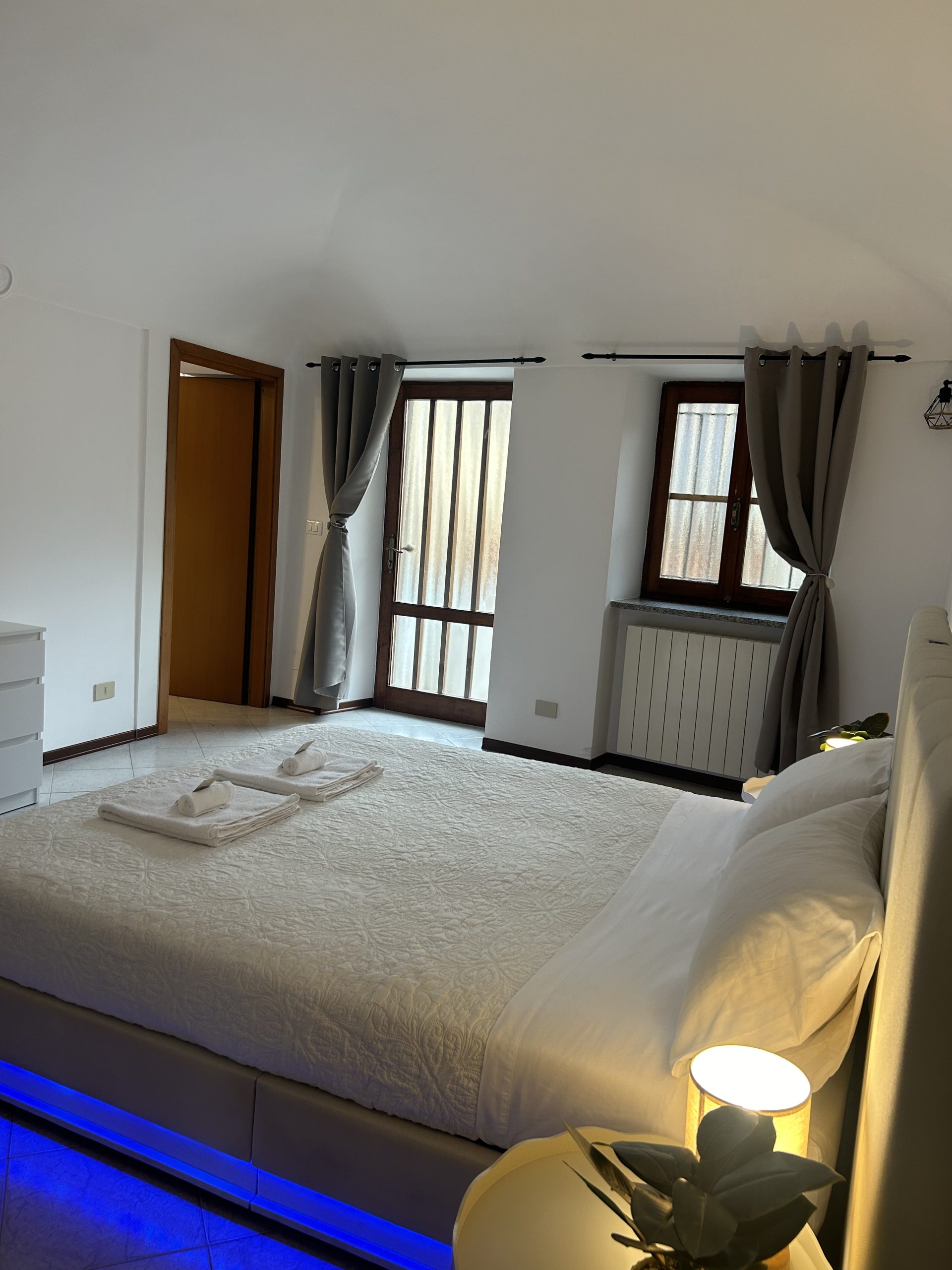 Una camera da letto luminosa con un grande letto ricoperto da una trapunta bianca, asciugamani piegati sopra, tende grigie alle finestre e alla porta, un comò bianco e luci LED blu sotto il letto. Sul comodino c'è una piccola lampada.
